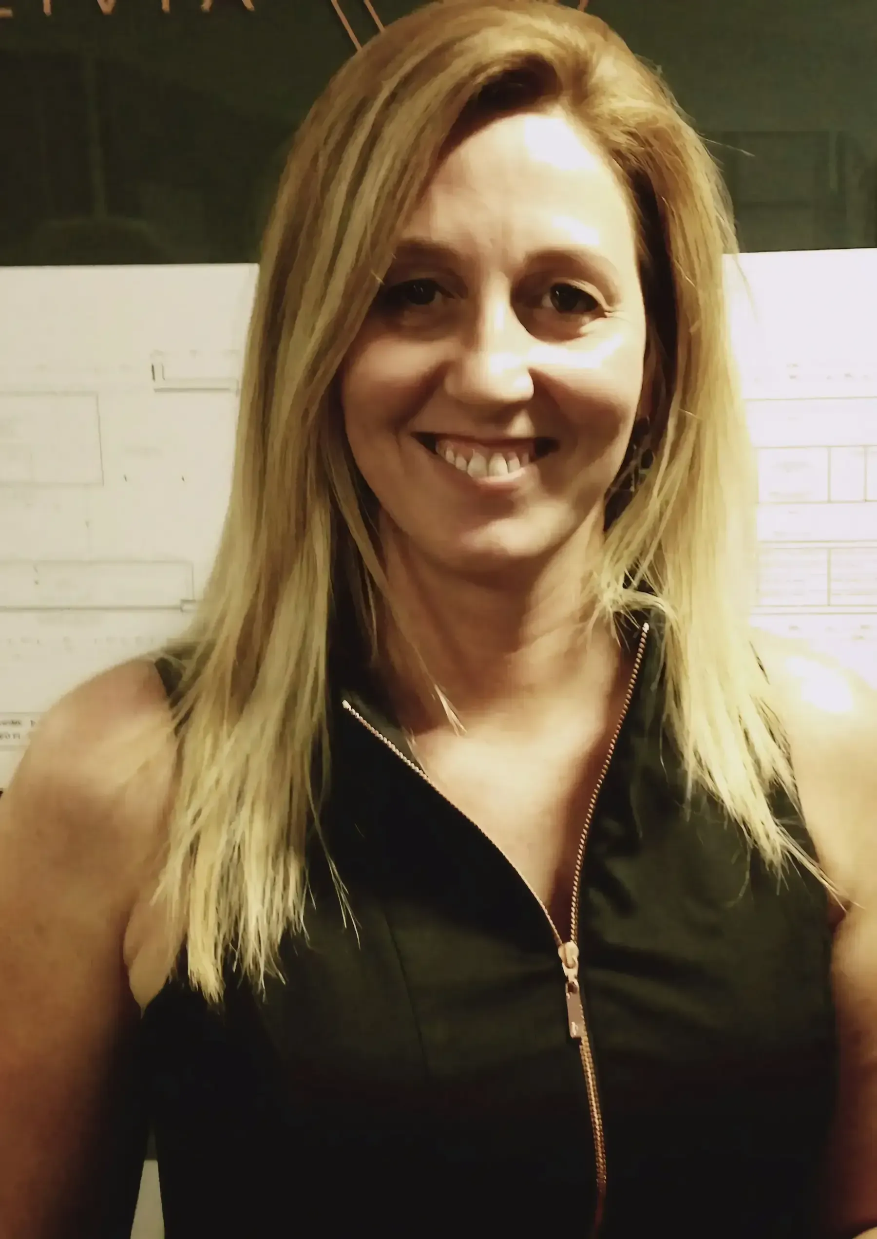 Smiling woman with blonde hair wearing a black sleeveless top against a white tiled wall.