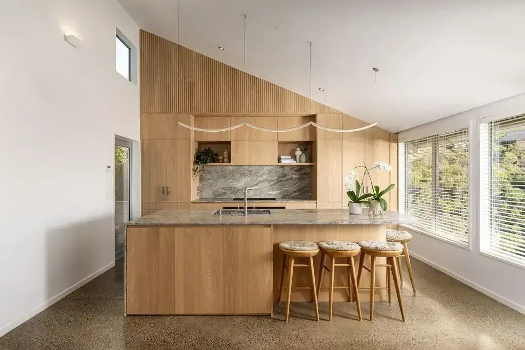 Modern kitchen with light wood cabinetry, stone countertops, and three stools.