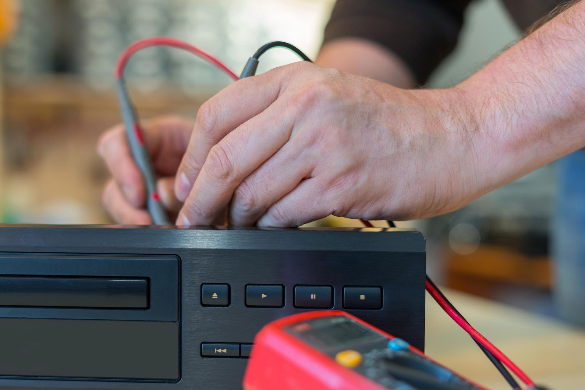 Hands testing wires on a black electronic device with a red multimeter in the foreground