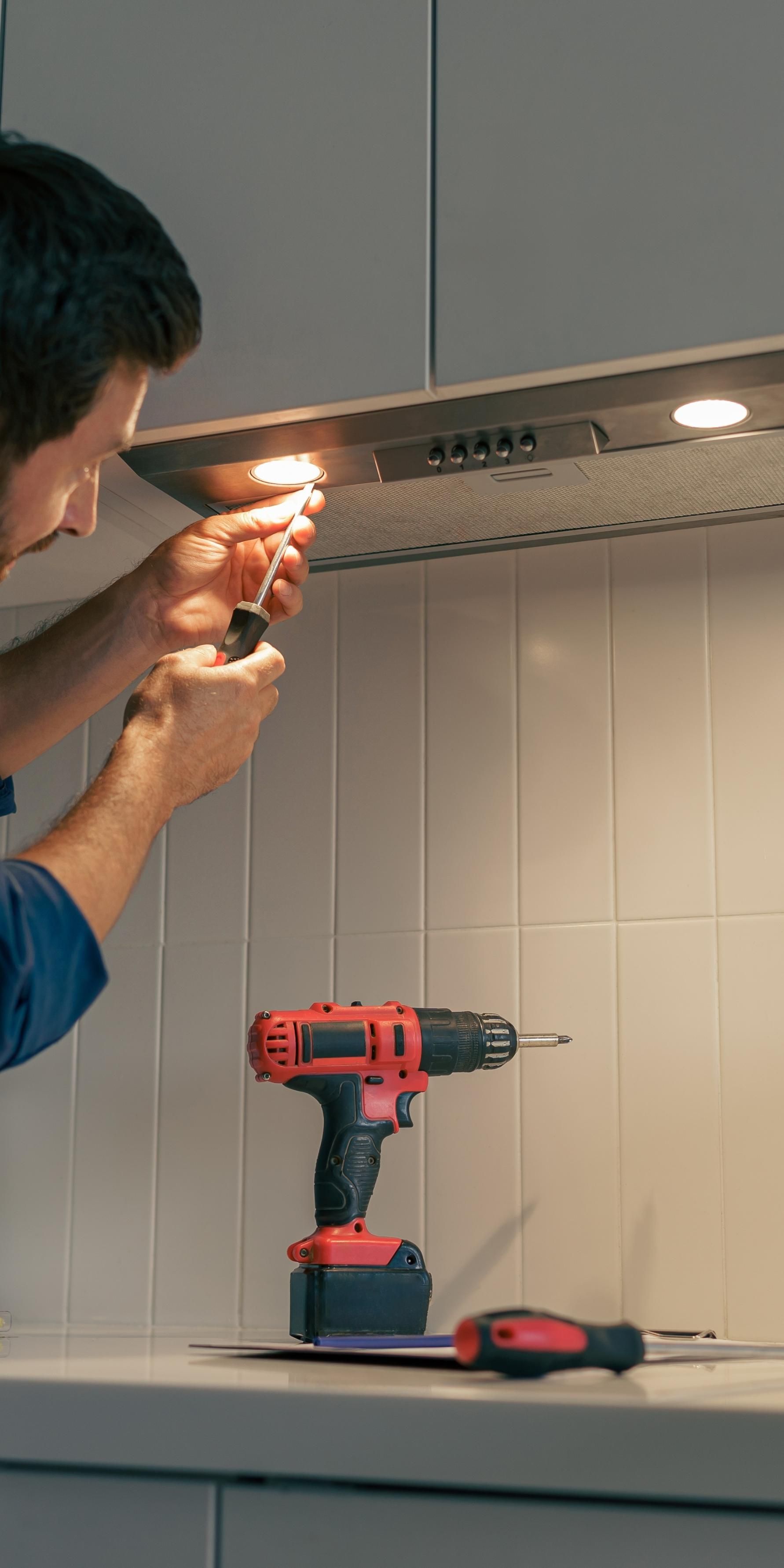 Person installing a kitchen under-cabinet light with a drill; red cordless drill on the counter below.