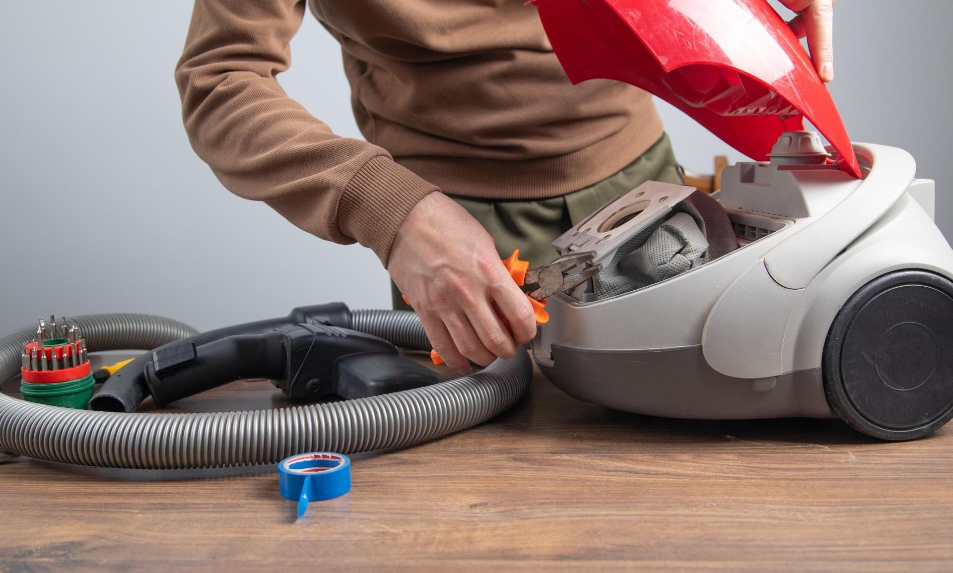 Person repairing a vacuum cleaner on a wooden table, with tools and the red cover open