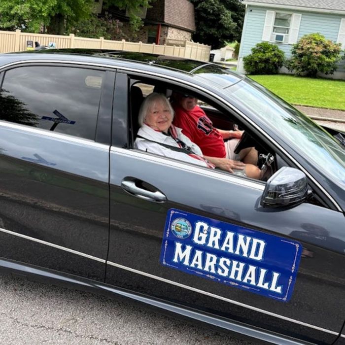 Gray sedan with “GRAND MARSHALL” sign on the door, two people inside, parked outdoors