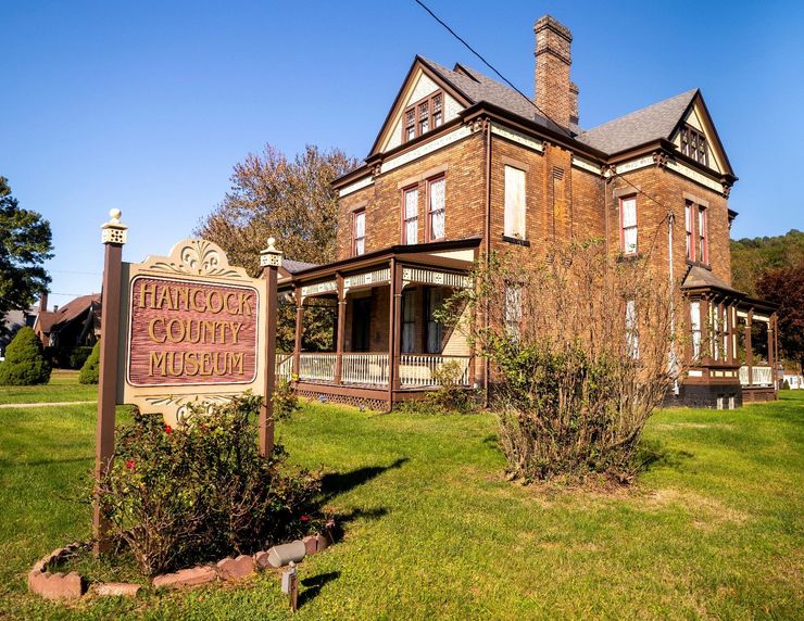 Historic brick house with porch and chimney beside a sign, surrounded by green lawn and trees.
