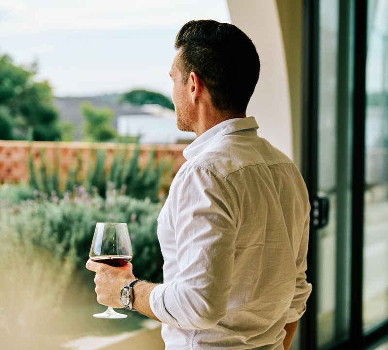 Stylish in a white shirt holding a wine glass, looking out a window at a garden view