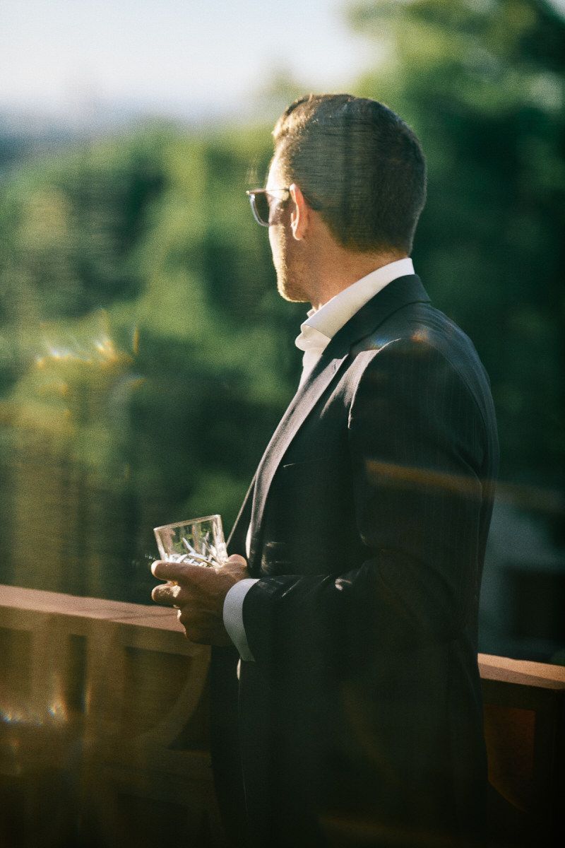 Sophisticated man in a dark suit holding a drink on a balcony, looking out over a blurred green landscape.