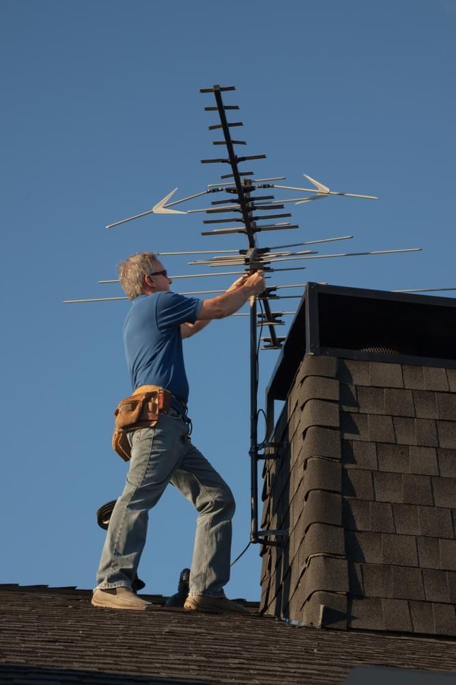 A Man Is Standing On Top Of A Roof Fixing An Antenna — Accent Electrics In Noraville, NSW
