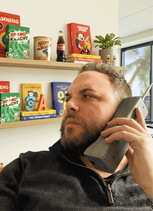 Een man houdt een grote, grijze mobiele telefoon aan zijn oor en kijkt met een serieuze uitdrukking opzij.