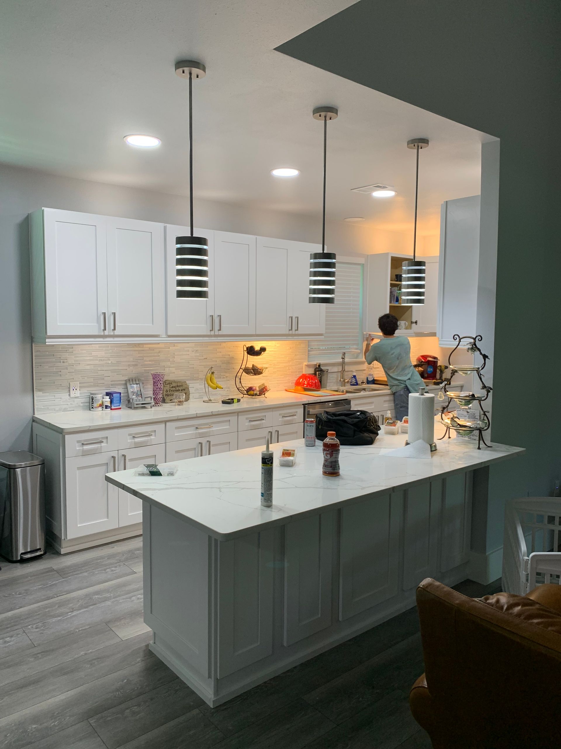 Bright modern kitchen with white cabinets, marble island, and a person cooking near the stove.