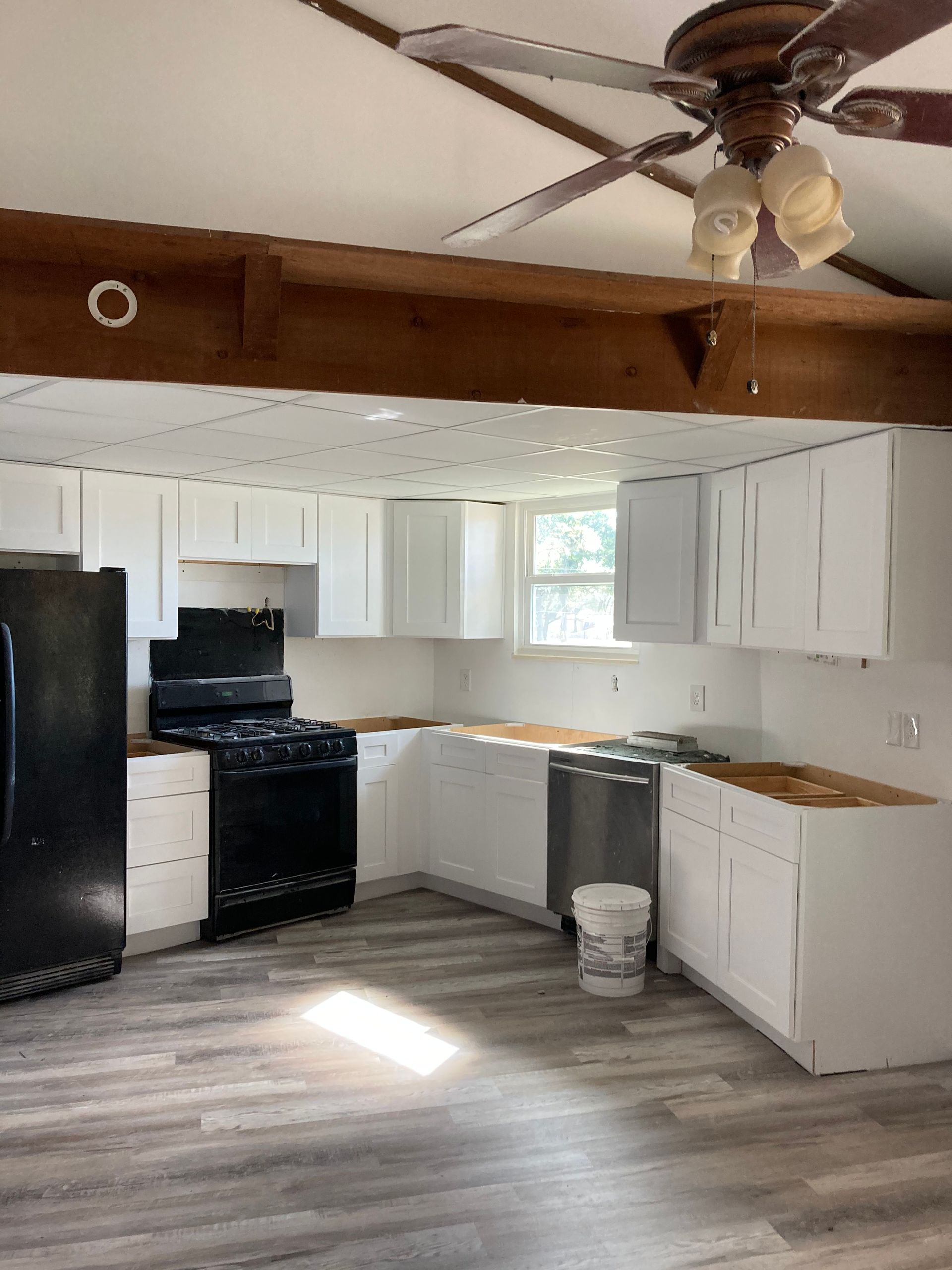 Small kitchen with white cabinets, black stove and fridge, exposed ceiling beam, and gray wood-look flooring.