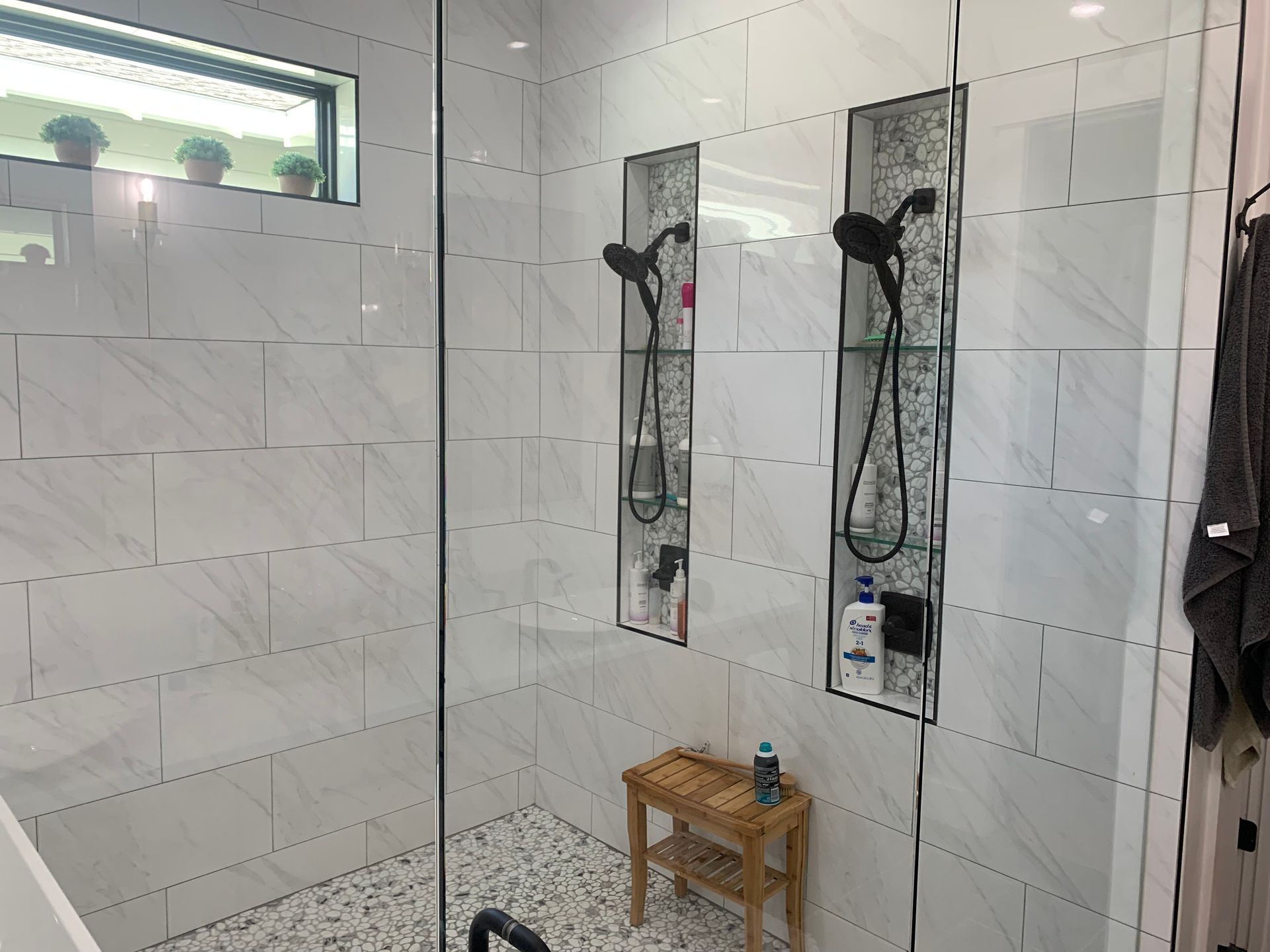 Walk-in shower with white tile, two black showerheads, a small wooden stool, and a window above.