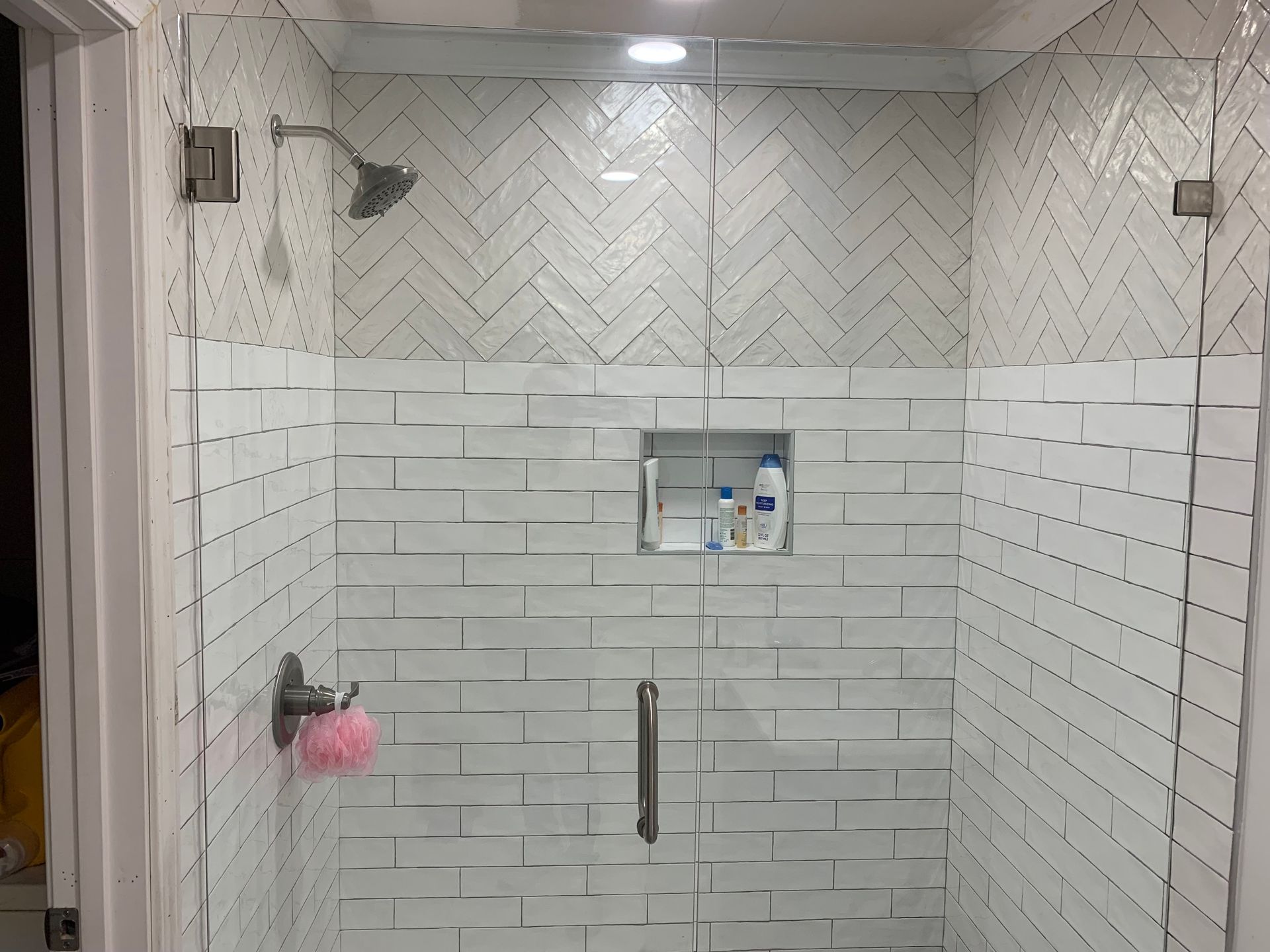 White tiled shower with glass enclosure, built-in shelf, and chrome handle, viewed from outside the stall
