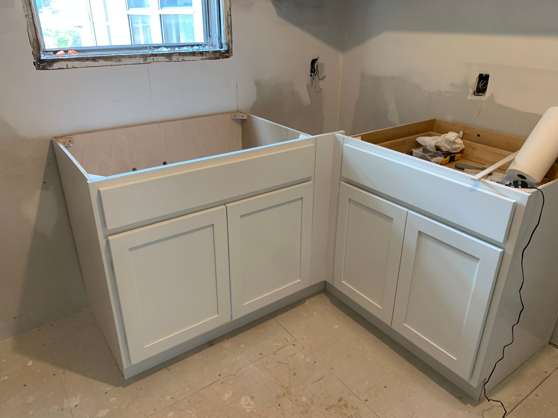 Partially installed white kitchen base cabinets in an unfinished room under a window