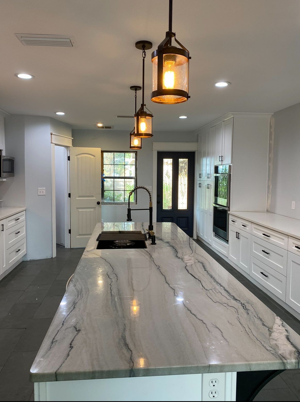 Modern kitchen with large marble island, black pendant lights, white cabinets, and double glass doors