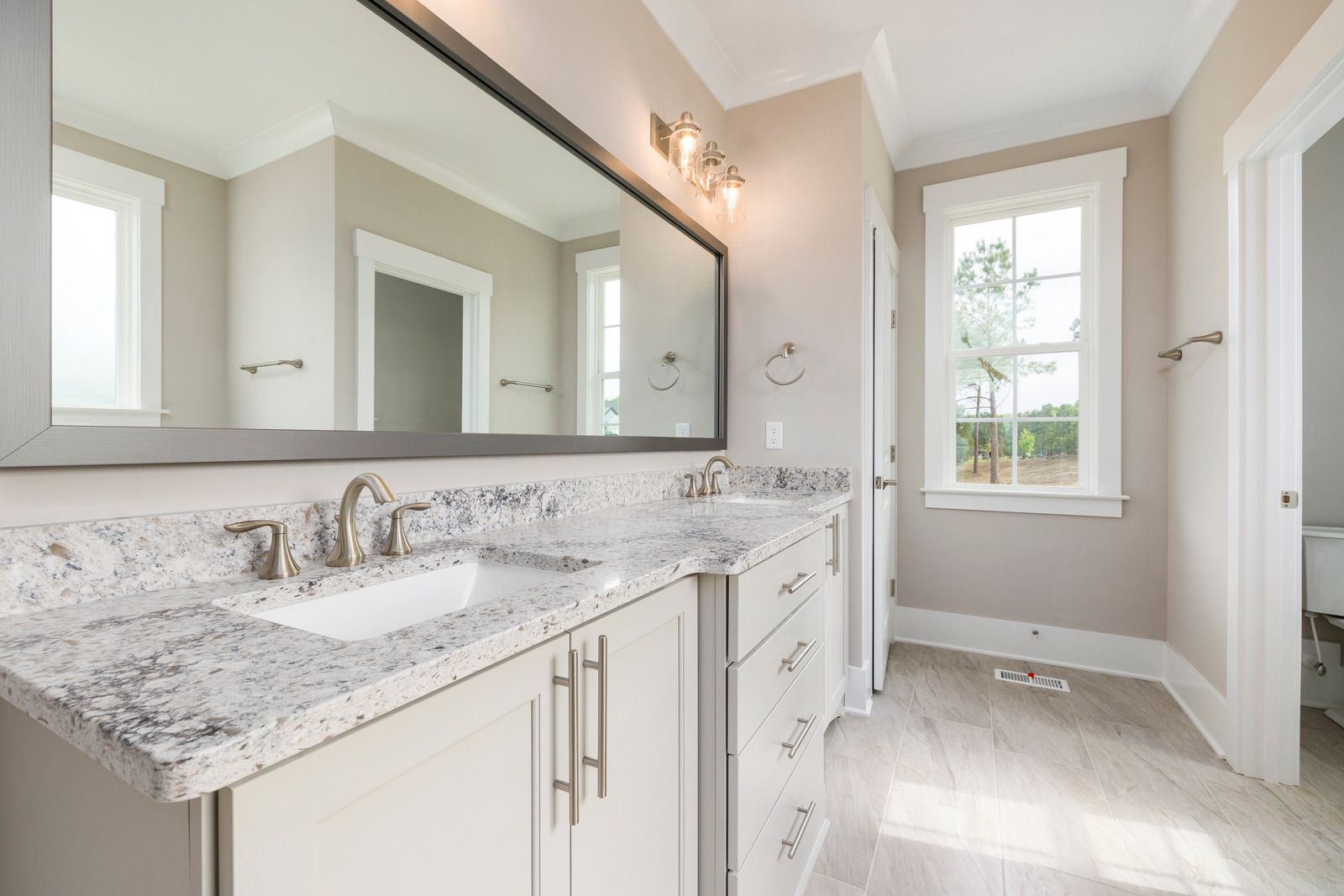 Bright bathroom with double vanity, large mirror, marble countertop, and window beside a shower.