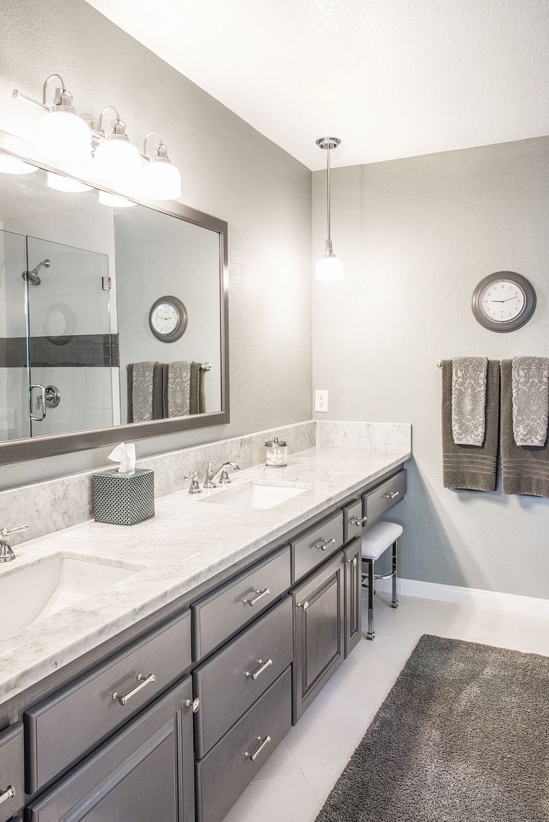 Modern bathroom with double vanity, large mirror, gray cabinets, wall sconces, and a patterned rug.