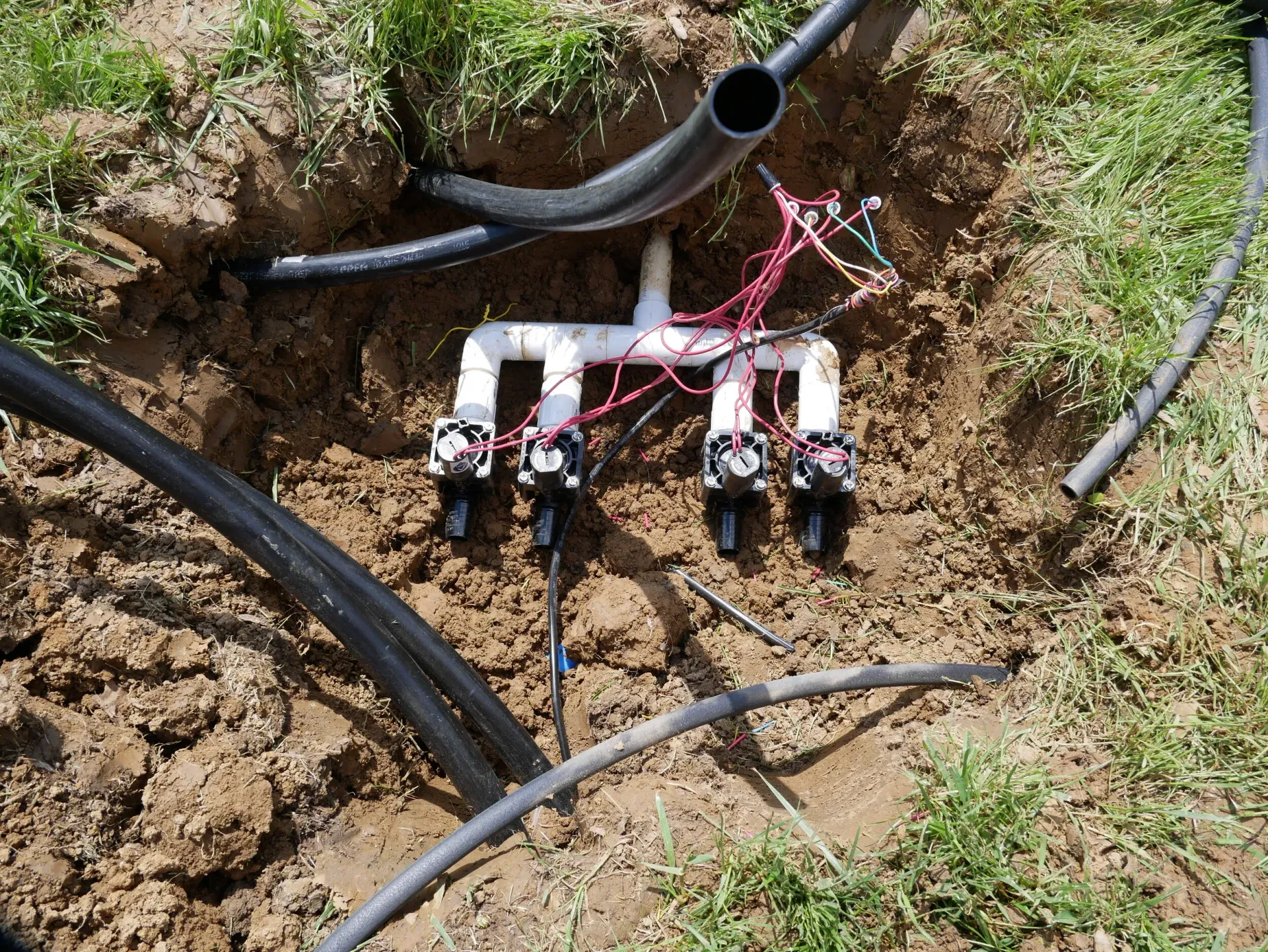 Broken irrigation pipe and exposed sprinkler manifold in a dirt trench with hoses and wires.