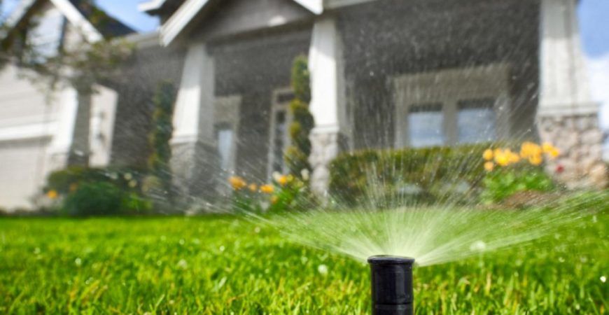 Sprinkler watering green lawn in front of a house with flowers and stone pillars