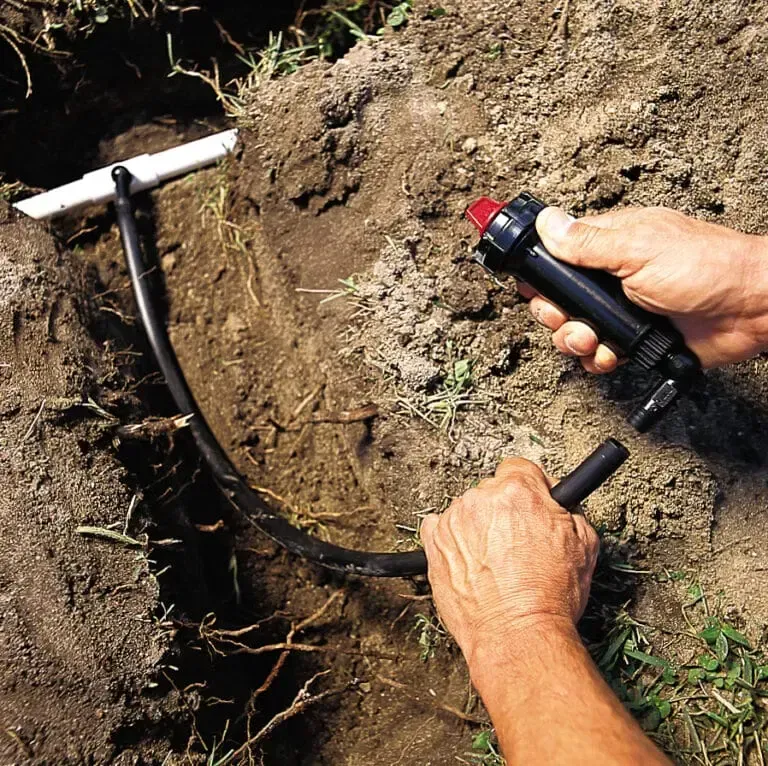 Hands using a tool to install or repair black tubing in a dirt trench outdoors