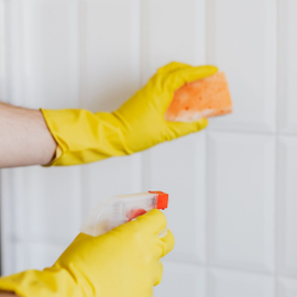 Gloved hands cleaning white tiles with an orange sponge and spray bottle