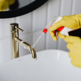 Yellow-gloved hands spraying bathroom sink faucet with cleaning product
