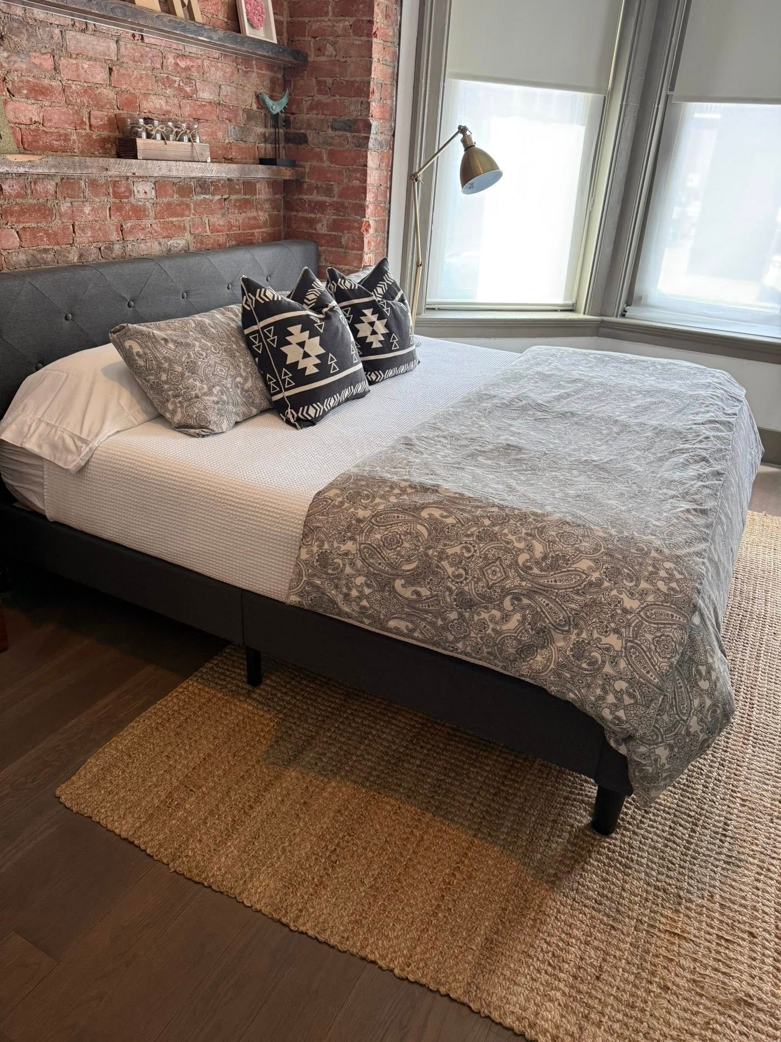 Bedroom with brick accent wall, bed with patterned bedding and pillows, and a window beside it.