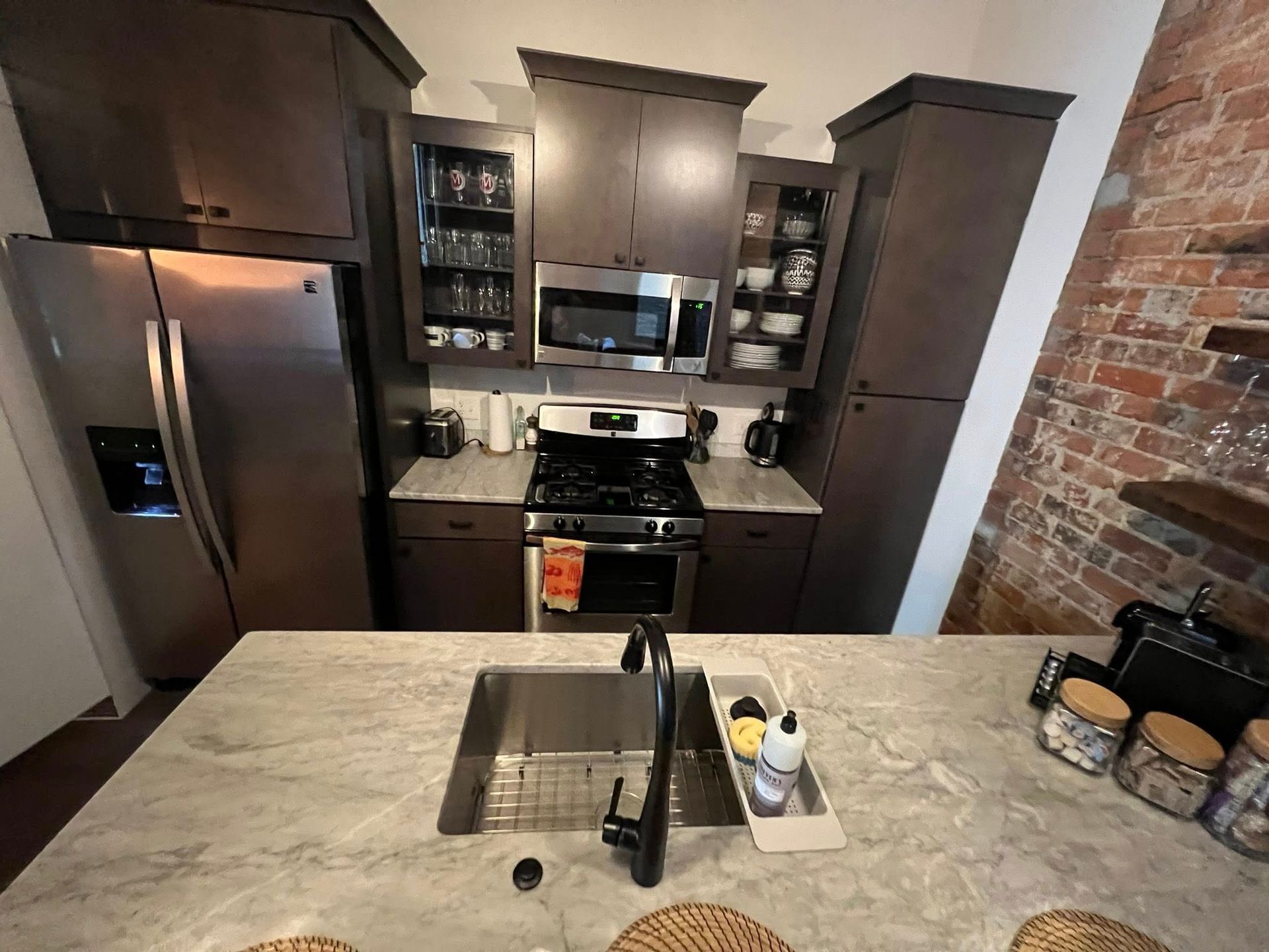 Modern kitchen with stainless steel appliances, dark cabinets, brick wall, and island sink with faucet
