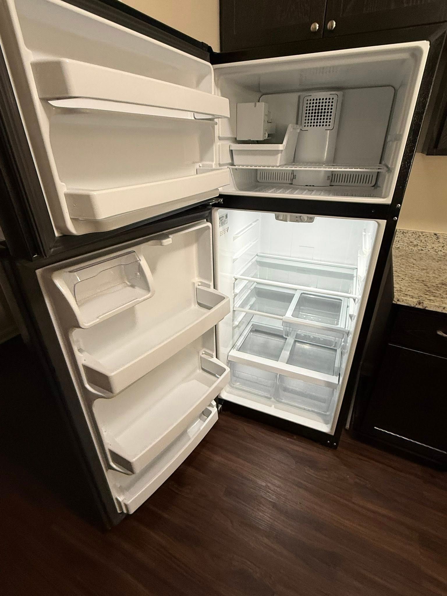 Open refrigerator with empty white shelves and door bins, viewed in a kitchen with dark wood floor