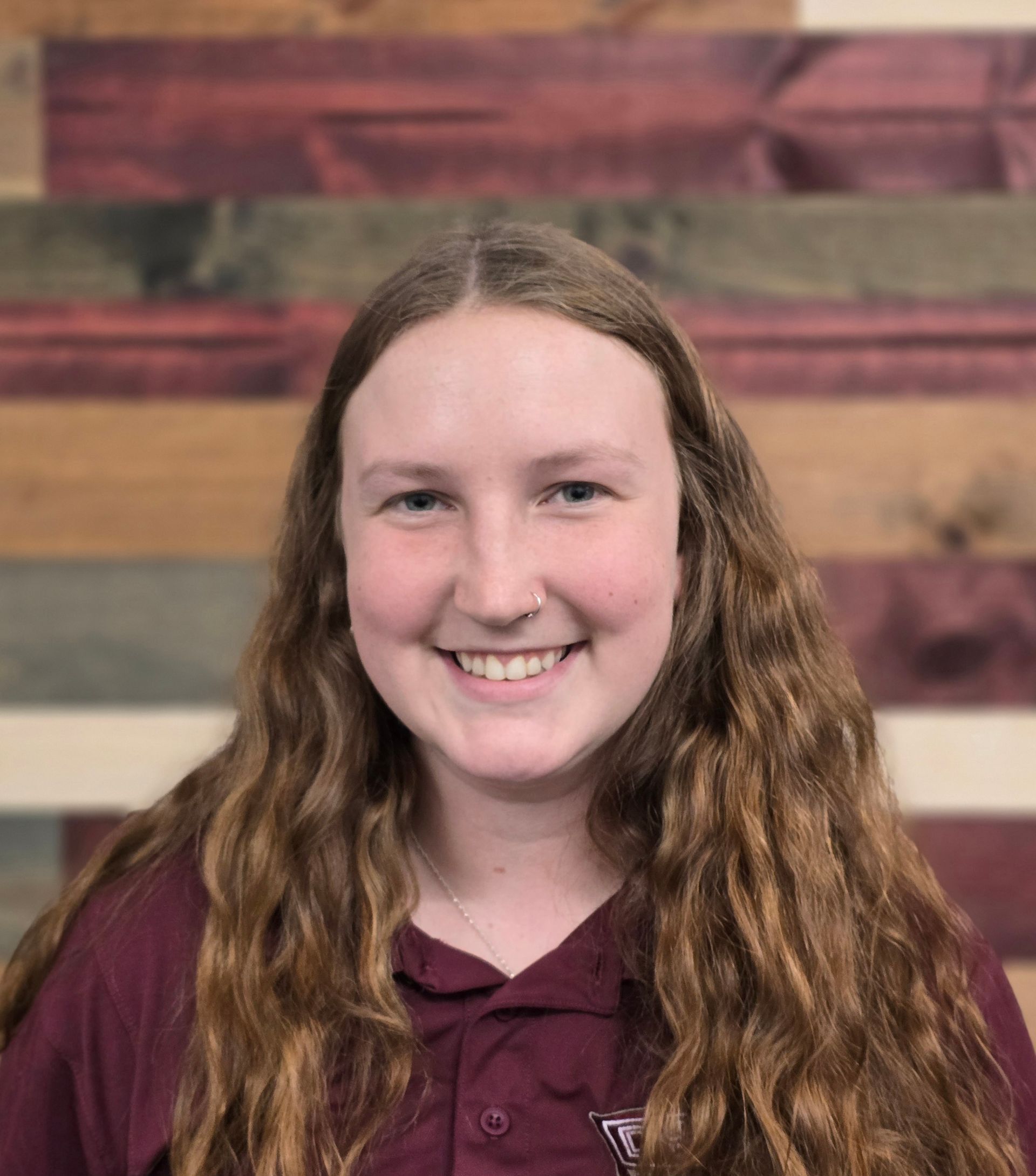 Young woman with long wavy brown hair, smiling, wearing maroon polo shirt, wooden backdrop.