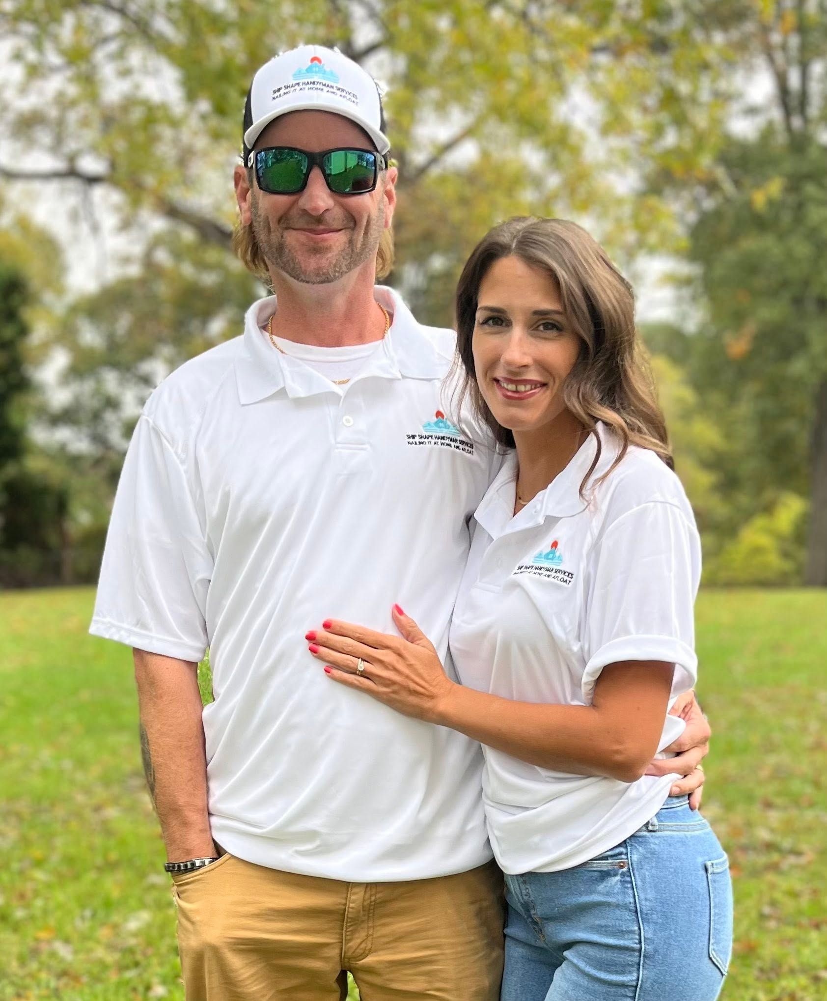 Two people in white polo shirts standing together outdoors on a grassy field, smiling at the camera.