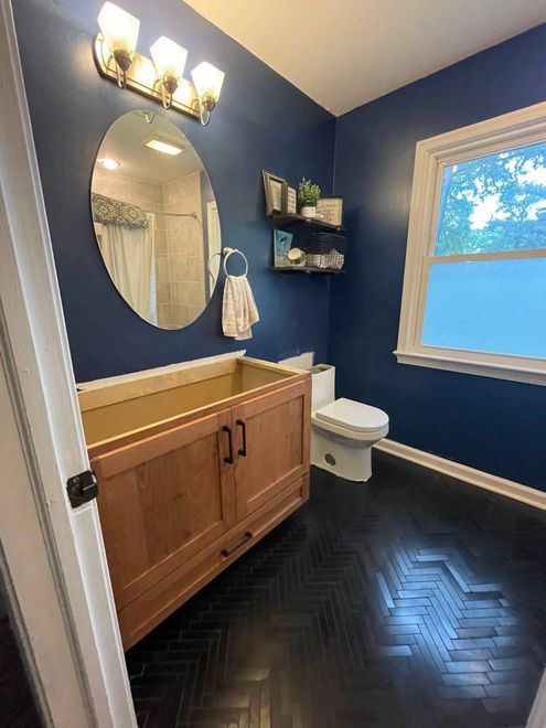 Blue bathroom with wood vanity, oval mirror, toilet, window, and dark herringbone floor.