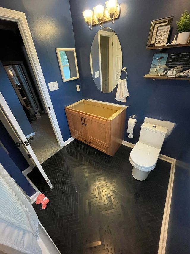 Blue bathroom with black herringbone floor, wood vanity, oval mirror, toilet, and wall shelf with decor.