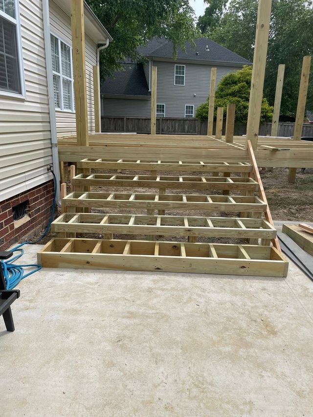 Unfinished wooden ramp framing beside a house, leading from a concrete driveway to a raised back deck.