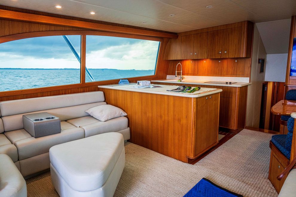Bright yacht cabin with tan sectional, white chair, wood cabinets, and ocean view through large windows