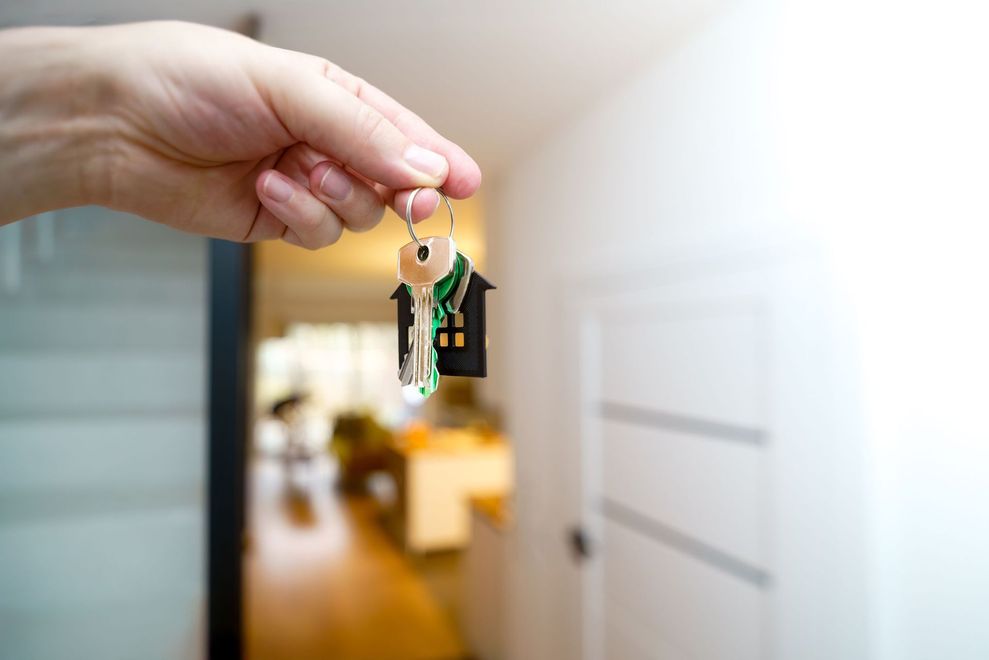 Hand holding house keys in front of a blurred apartment hallway, with a door in the background