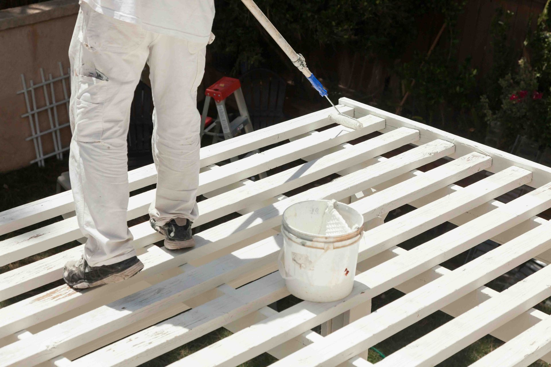 A person standing and painting a commercial pergola white with a white uniform and long reach paint roller