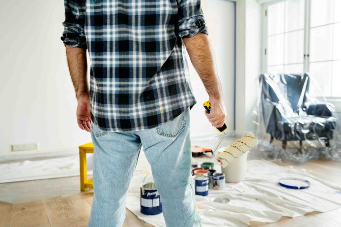 a man's plaid shirt from behind holding a paint roller in a white room with a covered chair in plastic