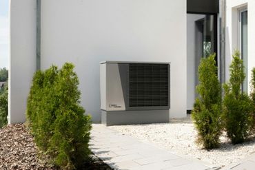 Heat pump unit next to a white building and small green bushes.