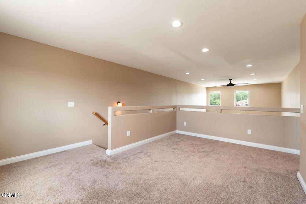 An empty carpeted loft area with light beige walls, recessed ceiling lights, and a wooden railing overlooking a lower floor.