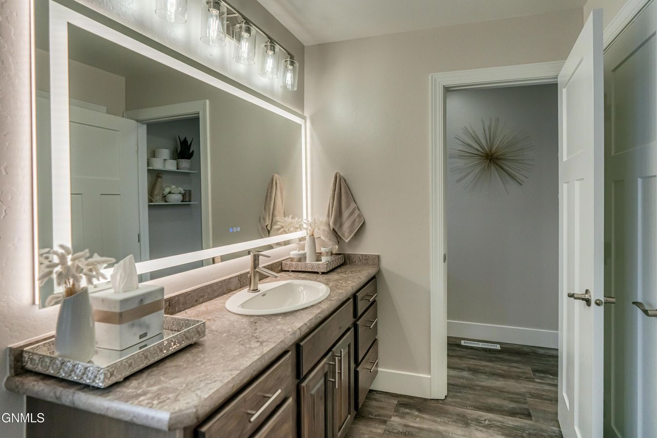 A bathroom with a vanity, sink, large mirror, and lighting, viewed through a doorway to an adjacent room.