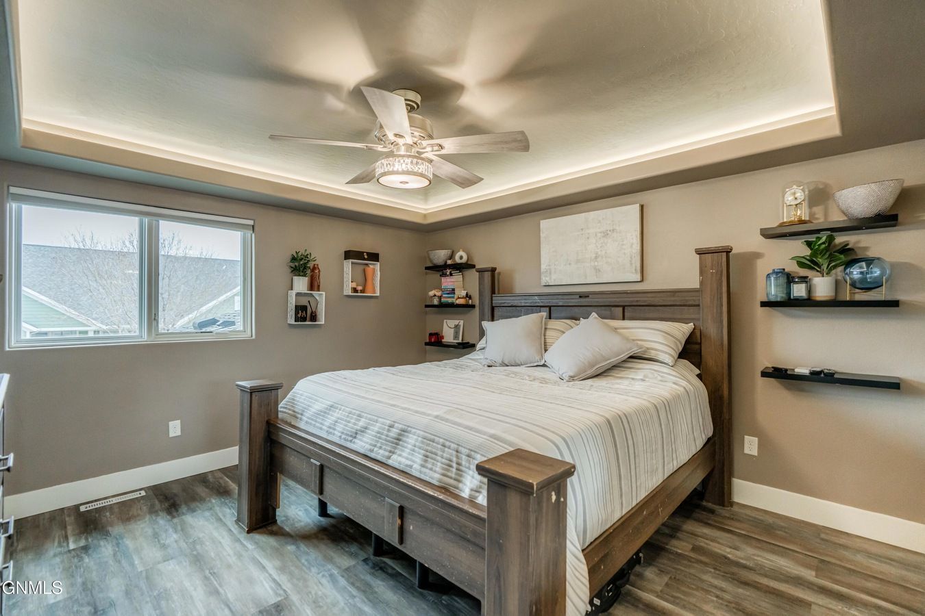 A bedroom with a wooden bed, gray walls, a tray ceiling with a fan, and floating shelves holding decor on both sides.
