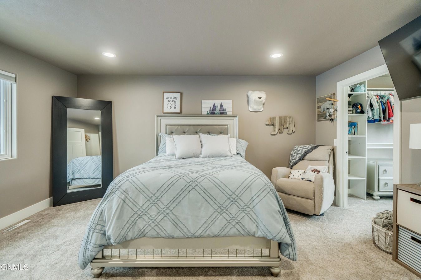 A bedroom with a queen bed featuring a blue patterned comforter, a large floor mirror, and a beige recliner near a closet.