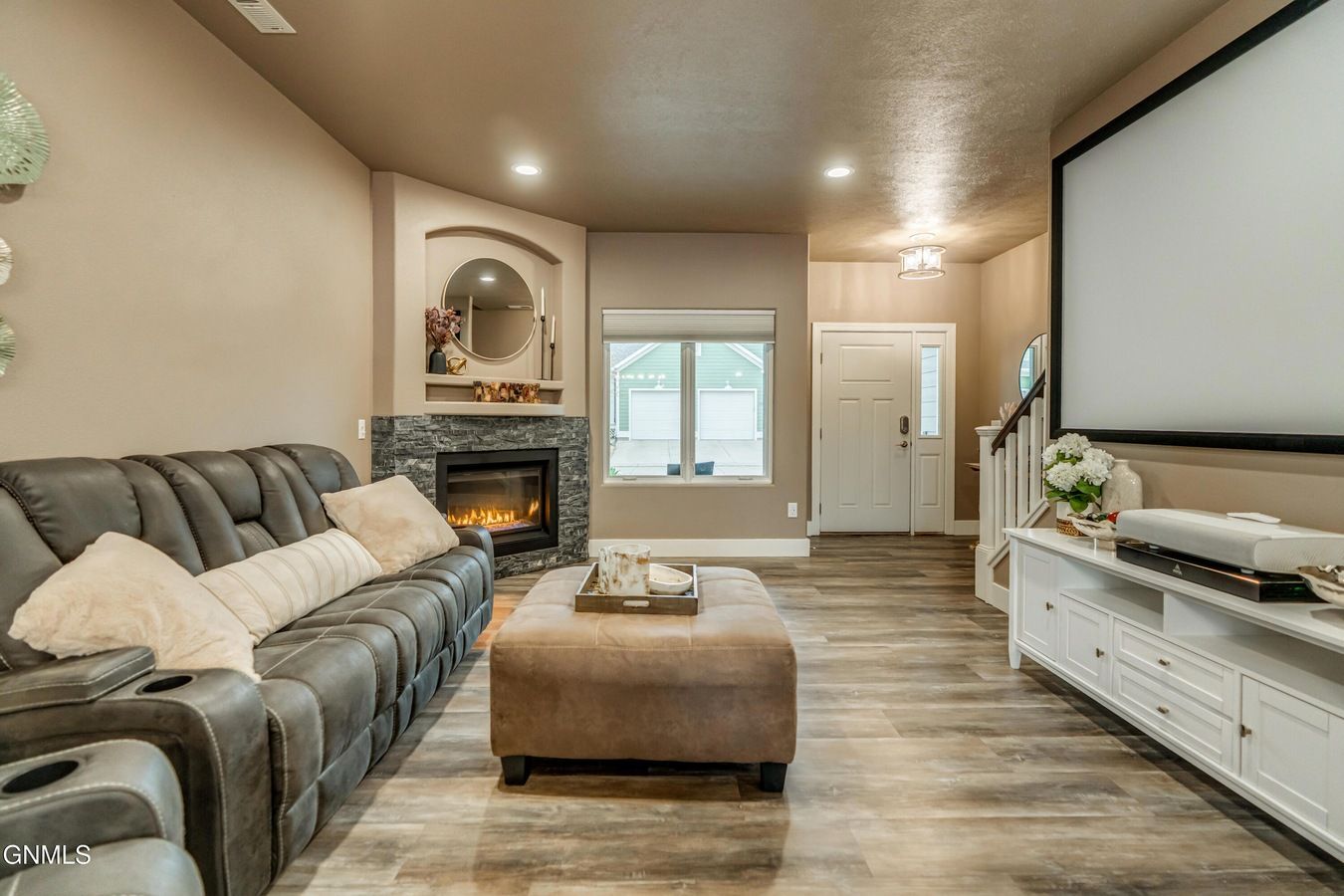 A basement living room with a grey reclining sofa, a tan ottoman, a fireplace with a stone surround, and a projector screen.