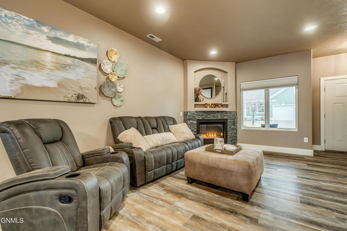 A gray recliner and sofa face a corner fireplace with a mounted mirror in a room with light walls and wood-look flooring.