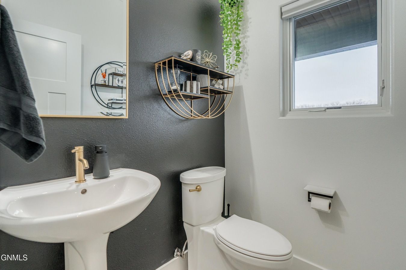 A modern half-bathroom with a pedestal sink, a dark grey accent wall, a gold-toned mirror and shelf, and a white toilet.