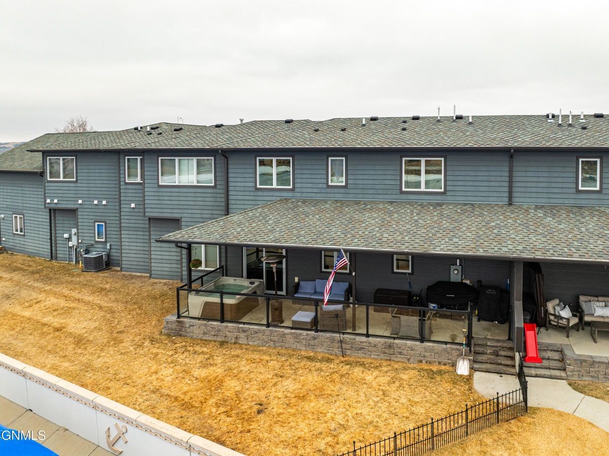 A multi-story gray home exterior featuring a covered patio, a hot tub, and outdoor seating over a dry, grassy yard.