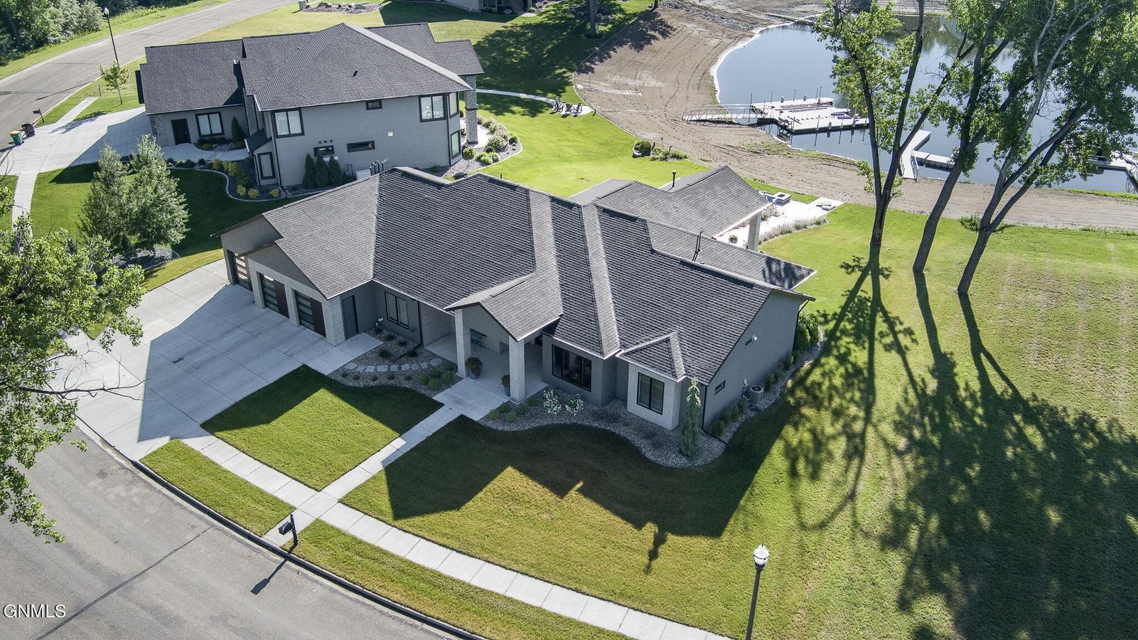 An aerial view of a gray suburban house with a dark shingled roof, a paved driveway, and a backyard facing a lake.