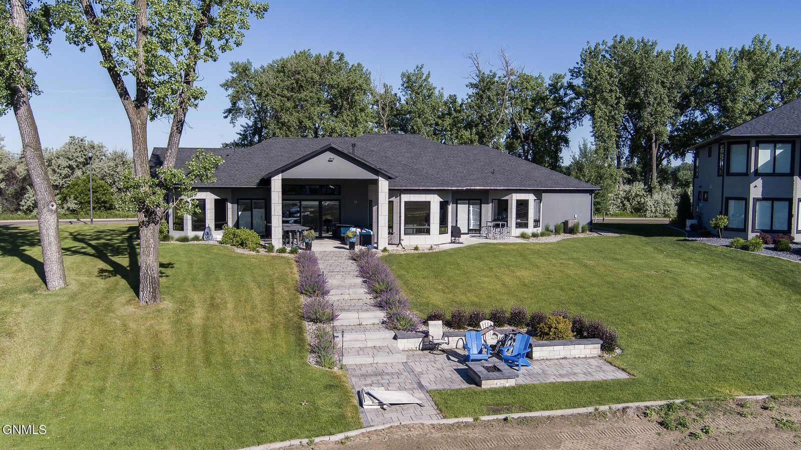 A dark-roofed suburban house with a stone pathway leading to a patio with fire pit chairs on a green lawn.