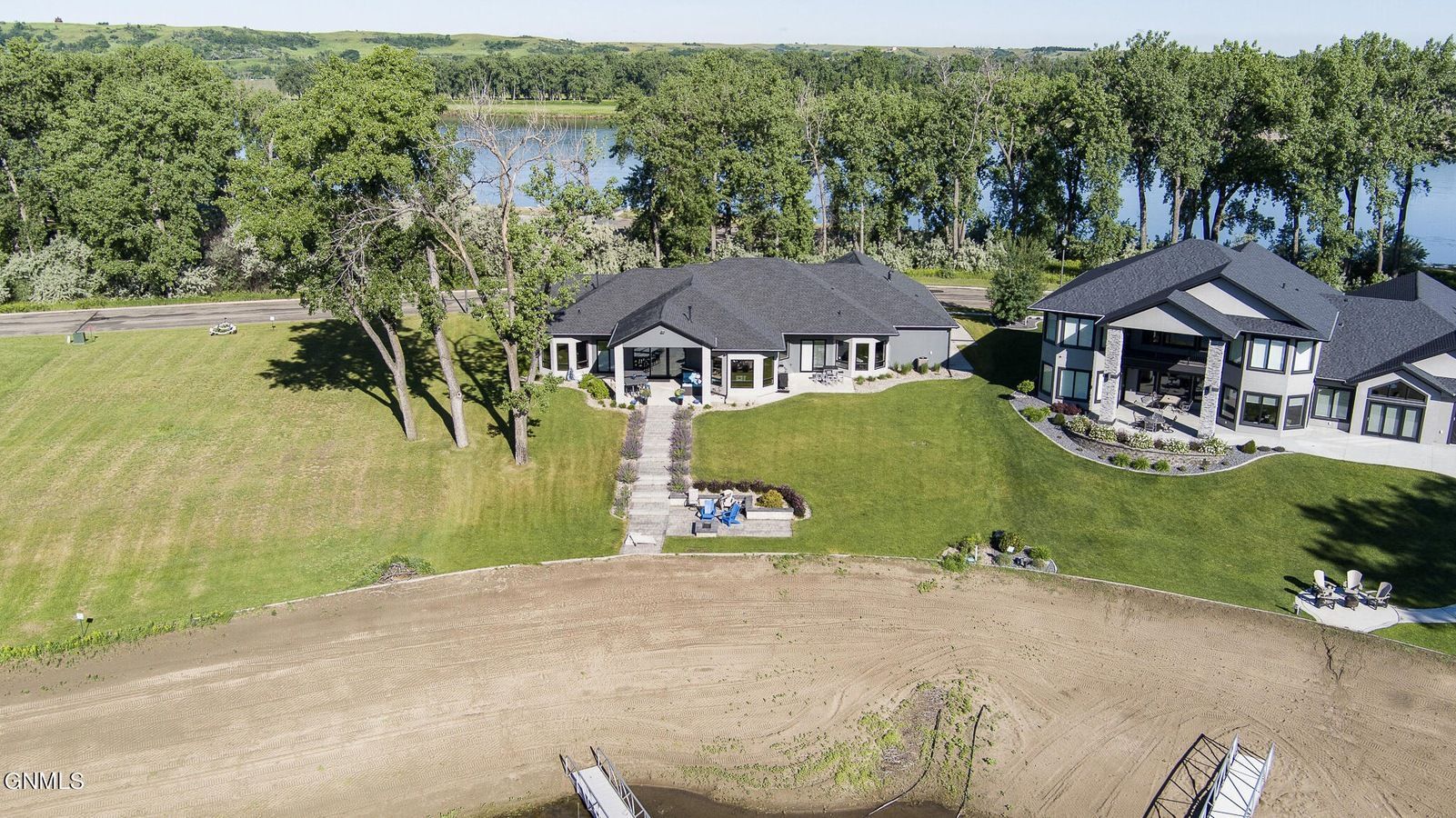 An aerial view of two large, modern gray houses on a grassy, sloped lot near a tree-lined lakefront with a dock.