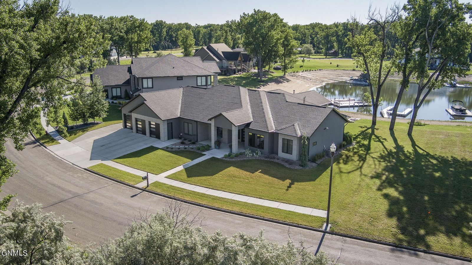 A modern, gray single-story house with a three-car garage and dark roof sits on a grassy lot next to a lake.