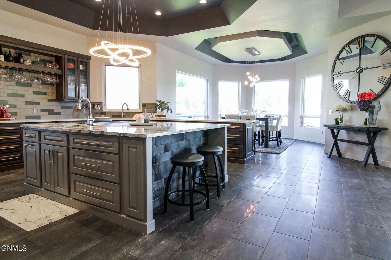 A modern kitchen featuring a dark grey center island with two bar stools, tiled floors, and a large decorative wall clock.
